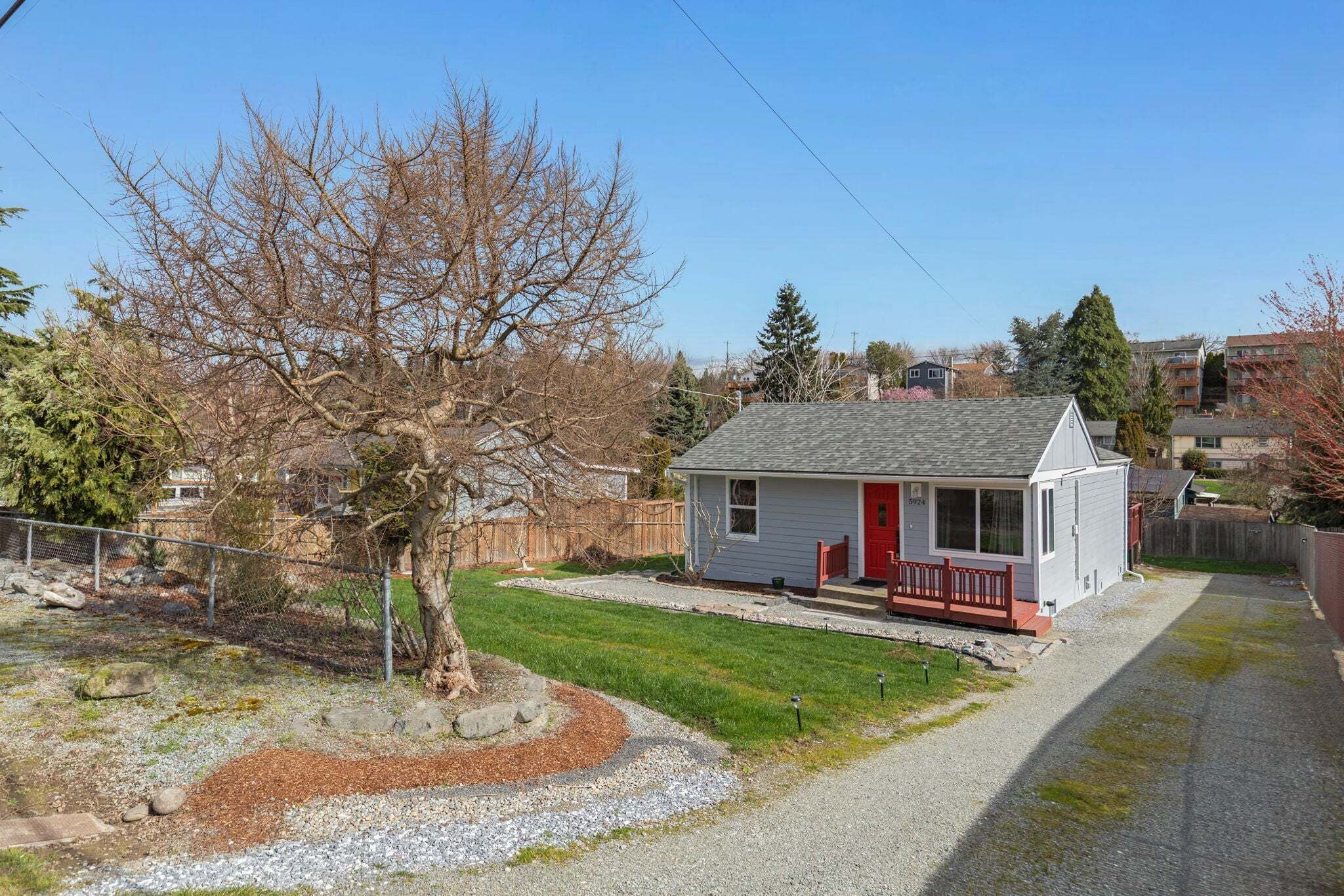 A small, single-story house with a red door and a porch, surrounded by a fenced yard and a gravel driveway.
