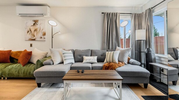 A modern living room with a grey sectional sofa, a wooden coffee table, and a large window with curtains. - Perch - AC Home w/ Sauna, Hot tub & Outdoor Seating! (Seattle)