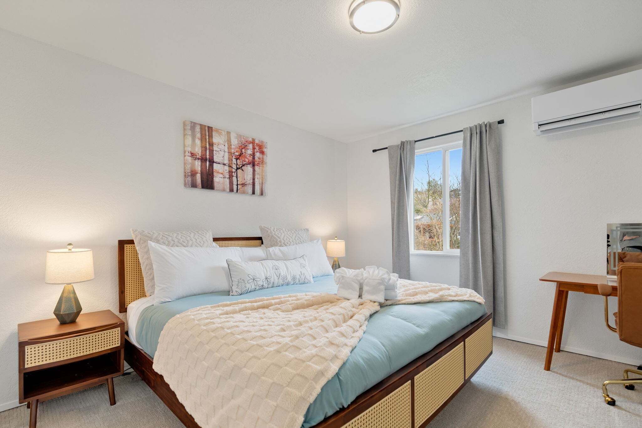 A bedroom with a bed, bedside tables, a desk, a window with curtains, and an air conditioning unit.