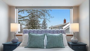 Ein Schlafzimmer mit großem Fenster, einem gepolsterten Kopfteil, zwei Nachttischen mit Lampen und einem Bett mit Kissen.