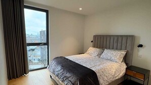 A modern bedroom with a large bed, a nightstand, a window with curtains, and a city view.