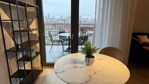 A modern living room with a round marble table, two cushioned chairs, a black metal shelving unit, and a view of a cityscape through a large sliding glass door.