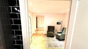 Un pasillo que conduce a una sala de estar con piso de madera, un sillón verde, un sofá gris y una lámpara de pie.