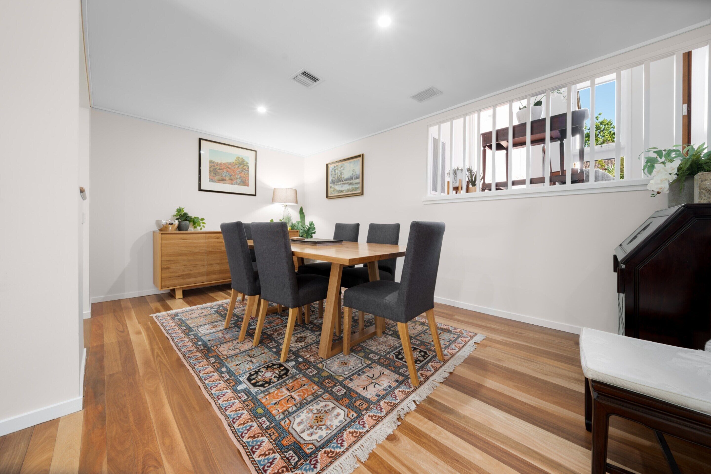 A dining area with a wooden table and chairs, a patterned rug, a wooden cabinet, and a window with blinds.