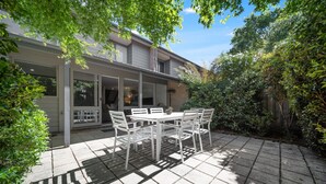 Un patio con tavolo e sedie, immerso nel verde e con una casa sullo sfondo.