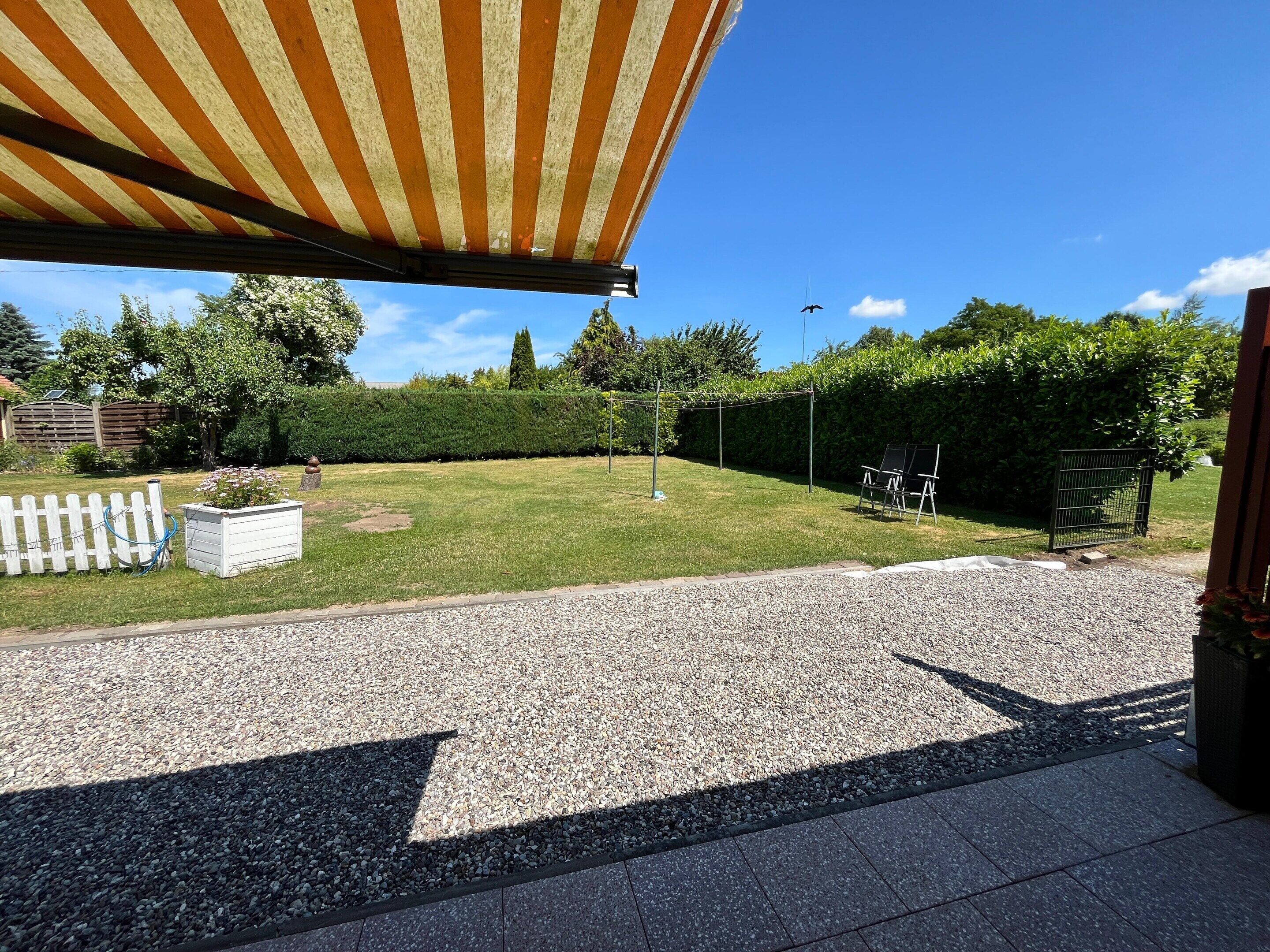 En patio med en iøjnefaldende markise, en grussti, en hæk og en indhegnet have.