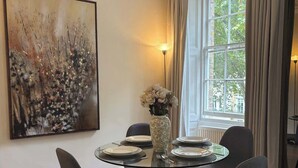 Un comedor con una mesa redonda de cristal, cuatro sillas, un jarrón con flores y un cuadro en la pared.