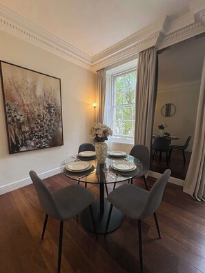 A dining area with a round glass table, four chairs, a vase with flowers, and a painting on the wall. - Stylish 1 Bed in Knightsbridge Prime Location (Greater London)