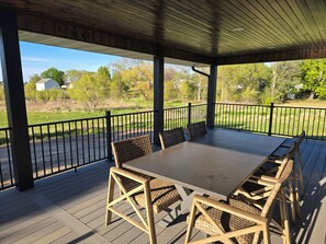Une terrasse extérieure couverte, dotée d’une table et de chaises, entourée d’une rambarde en métal noir, et offrant une vue sur une zone verdoyante avec des arbres et un bâtiment situé au loin.