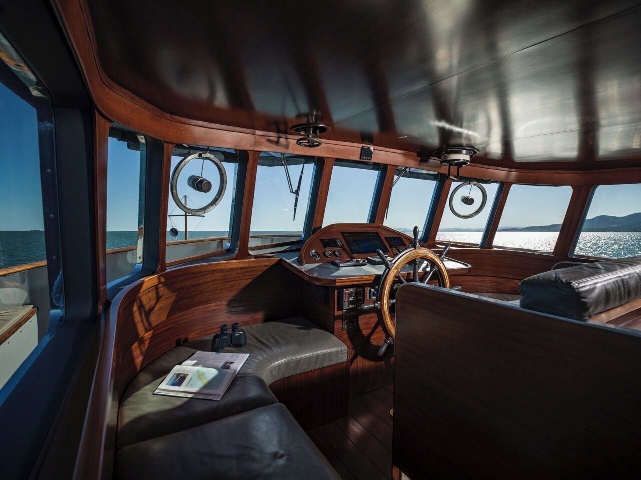 Interior de un barco con timón, cubierta de madera y grandes ventanas que ofrecen vistas al agua.