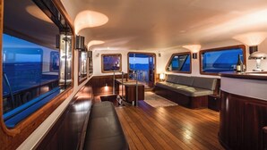 A yacht interior with a wooden deck, a bar area, seating, and large windows offering views.