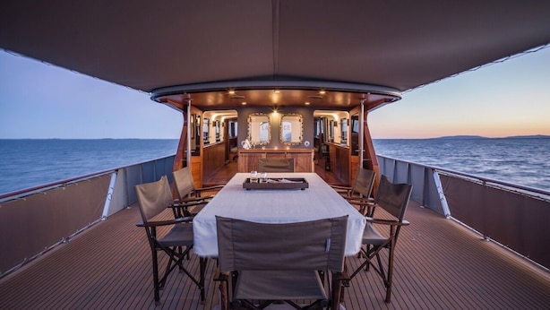 A dining area on a boat with a table set for four, surrounded by chairs, and a view of the sea.