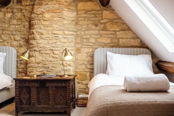 Ein Schlafzimmer mit Steinsturz, schrägen Decken, einem hölzernen Nachttisch mit Lampe, einem Bett mit aufgerolltem Handtuch und einem gestreiften Kissen.