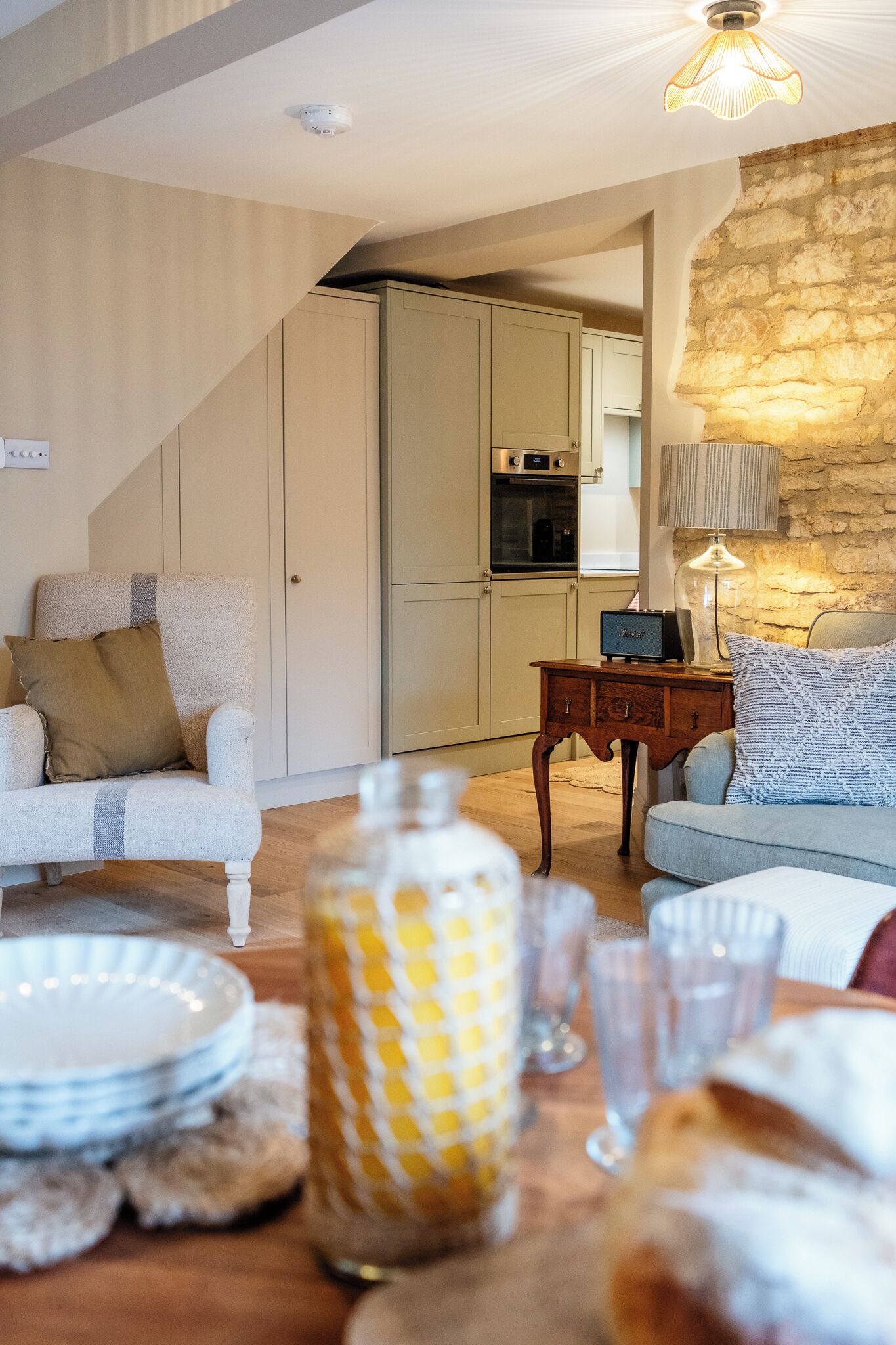 A living room with a sofa, a table with plates and glasses, a lamp, and a kitchen in the background.