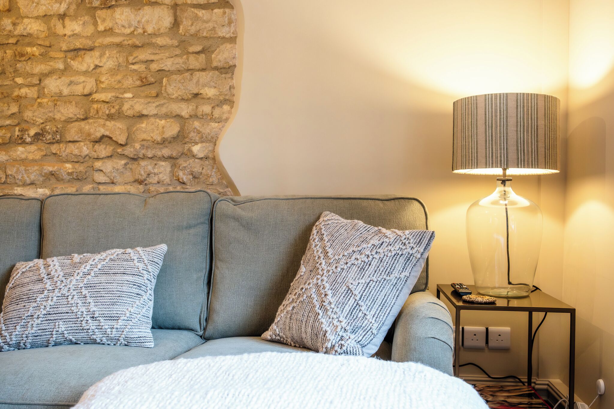 Una habitación con pared de piedra, un sofá gris con cojines estampados, una mesita de noche con una lámpara y un enchufe.