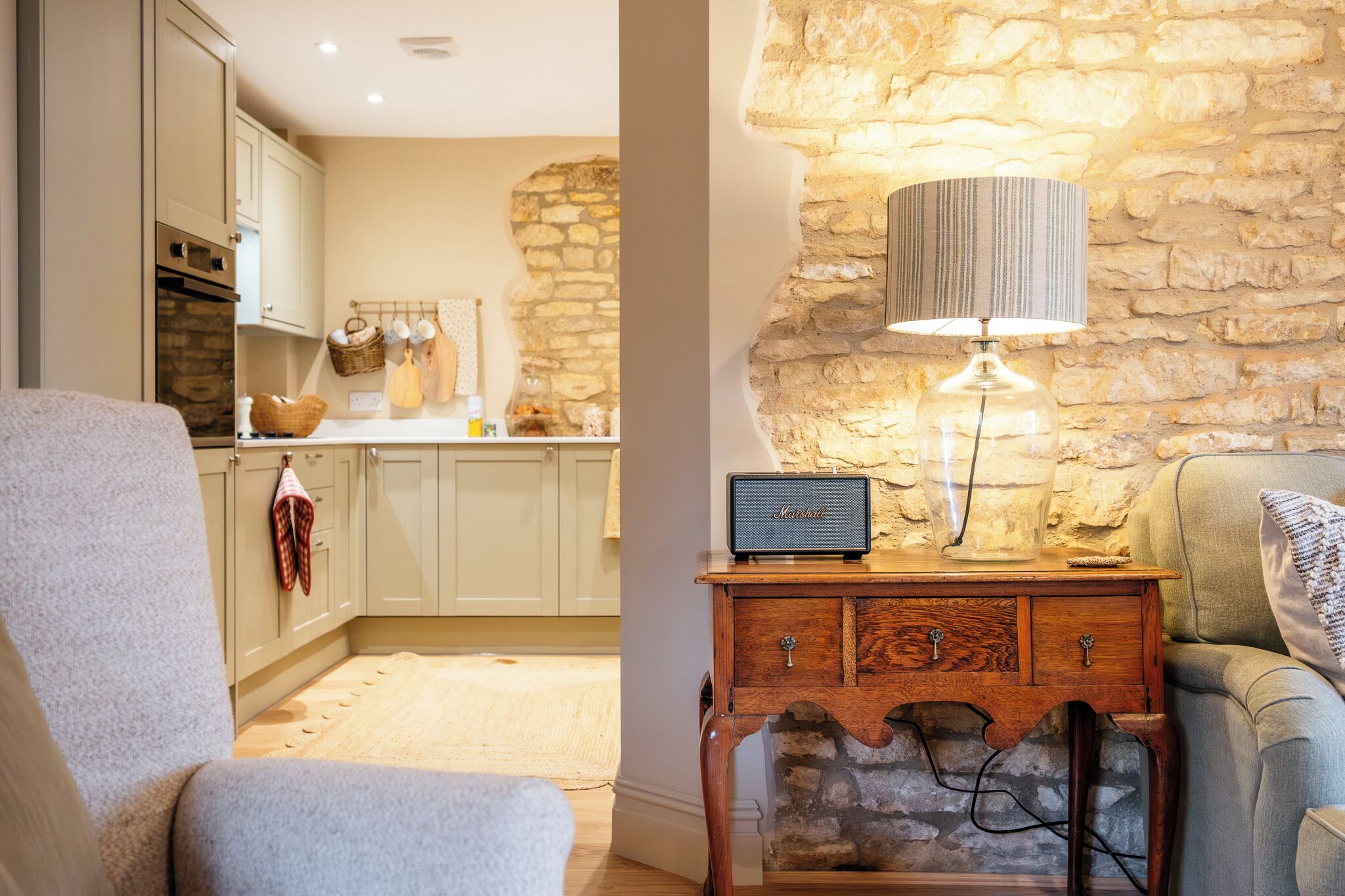 A cozy living room with a stone wall, a lamp, a wooden side table, and a kitchen in the background.