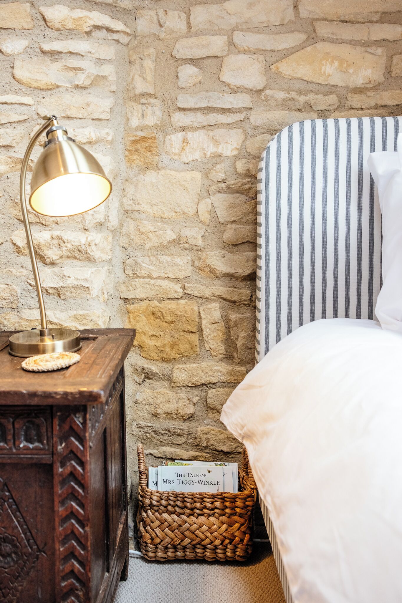 Una pared de piedra, una lámpara de noche, un cojín a rayas, una cesta con un libro y una mesita de noche de madera.