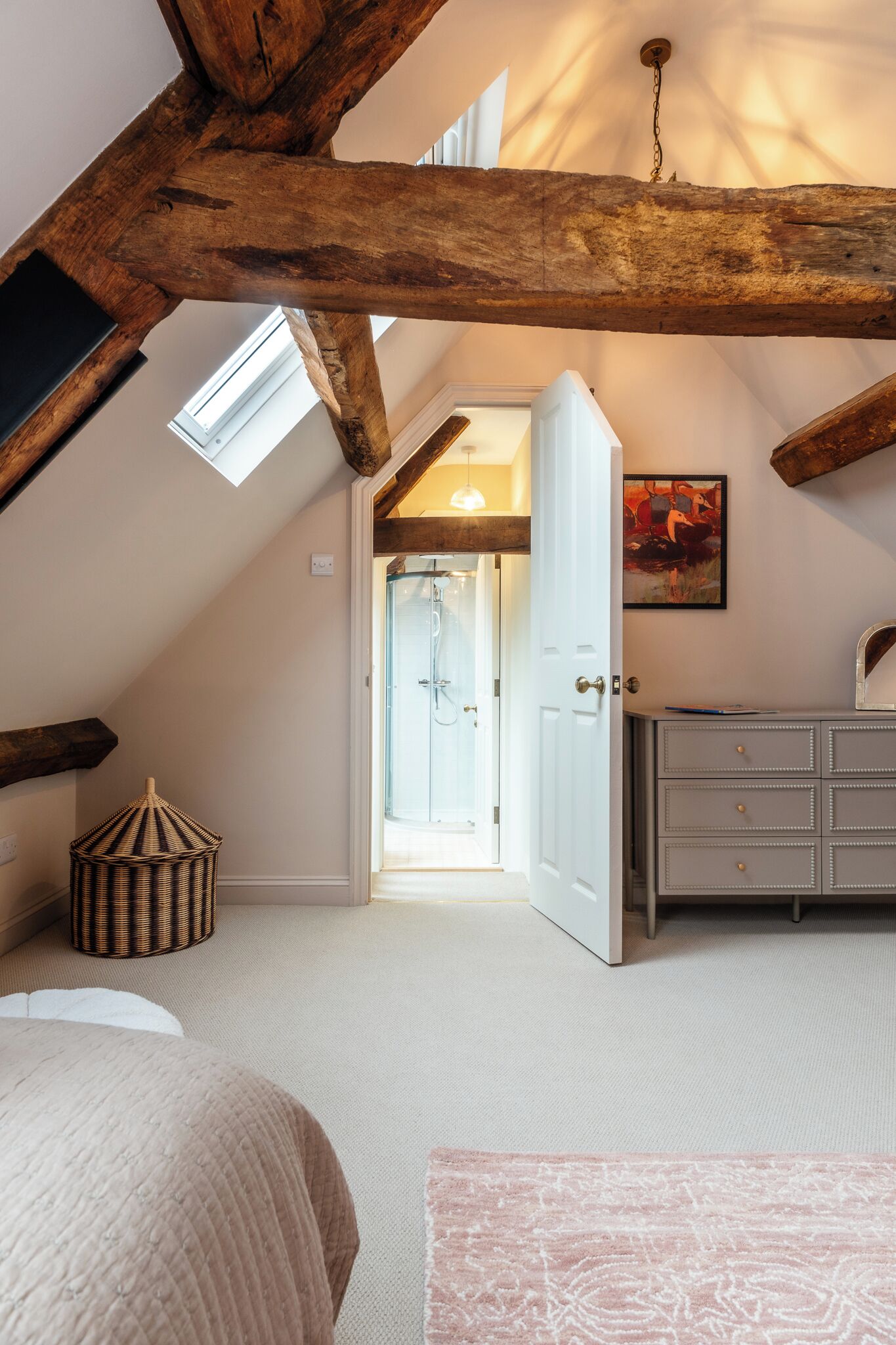 Een slaapkamer met een schuine kap, een bed, een dressoir, een rieten mand en een deur naar een badkamer.