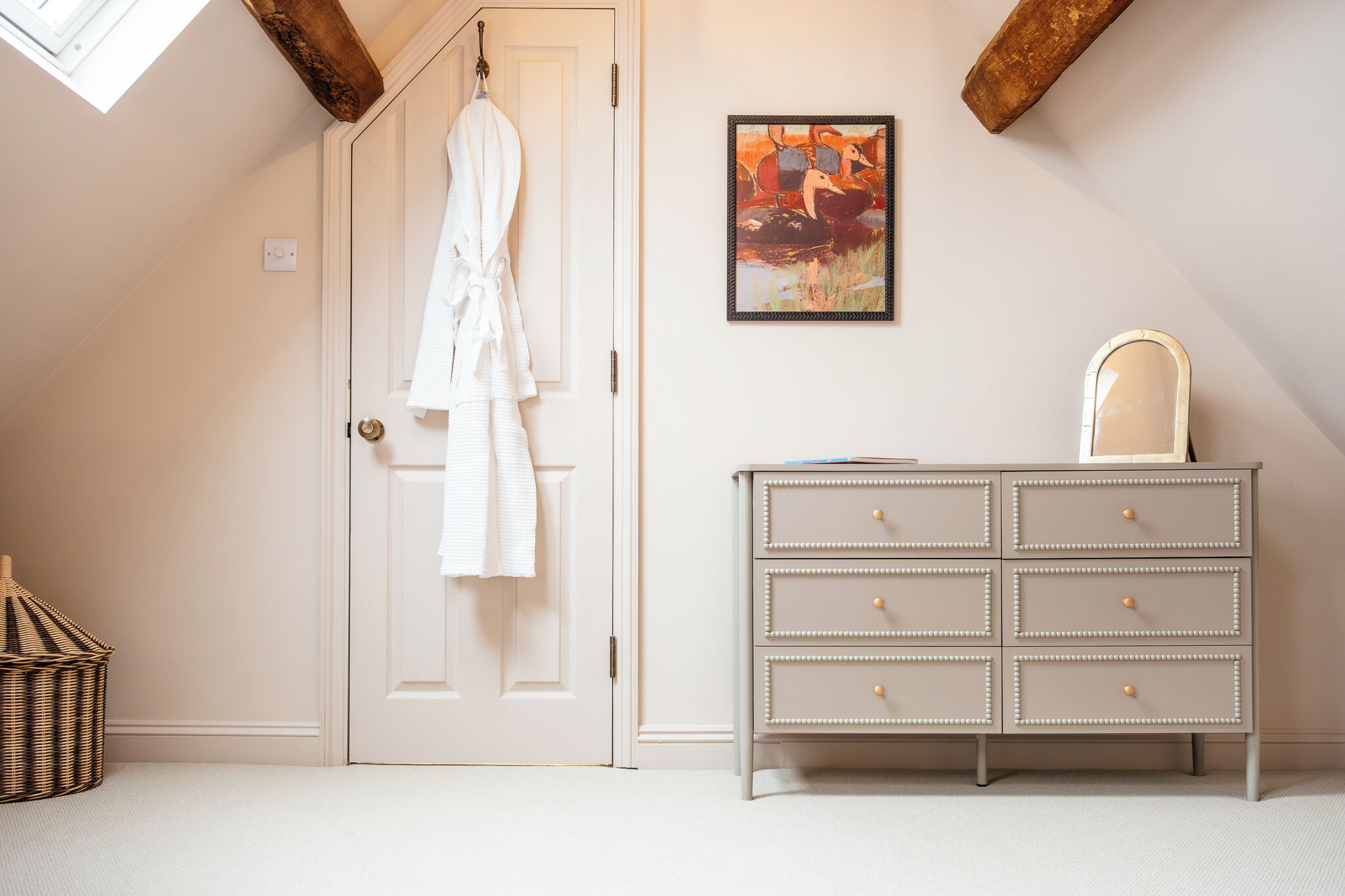 A room with a white door, a white robe hanging on the door, a dresser with drawers, a framed picture on the wall, and a wicker basket on the floor.