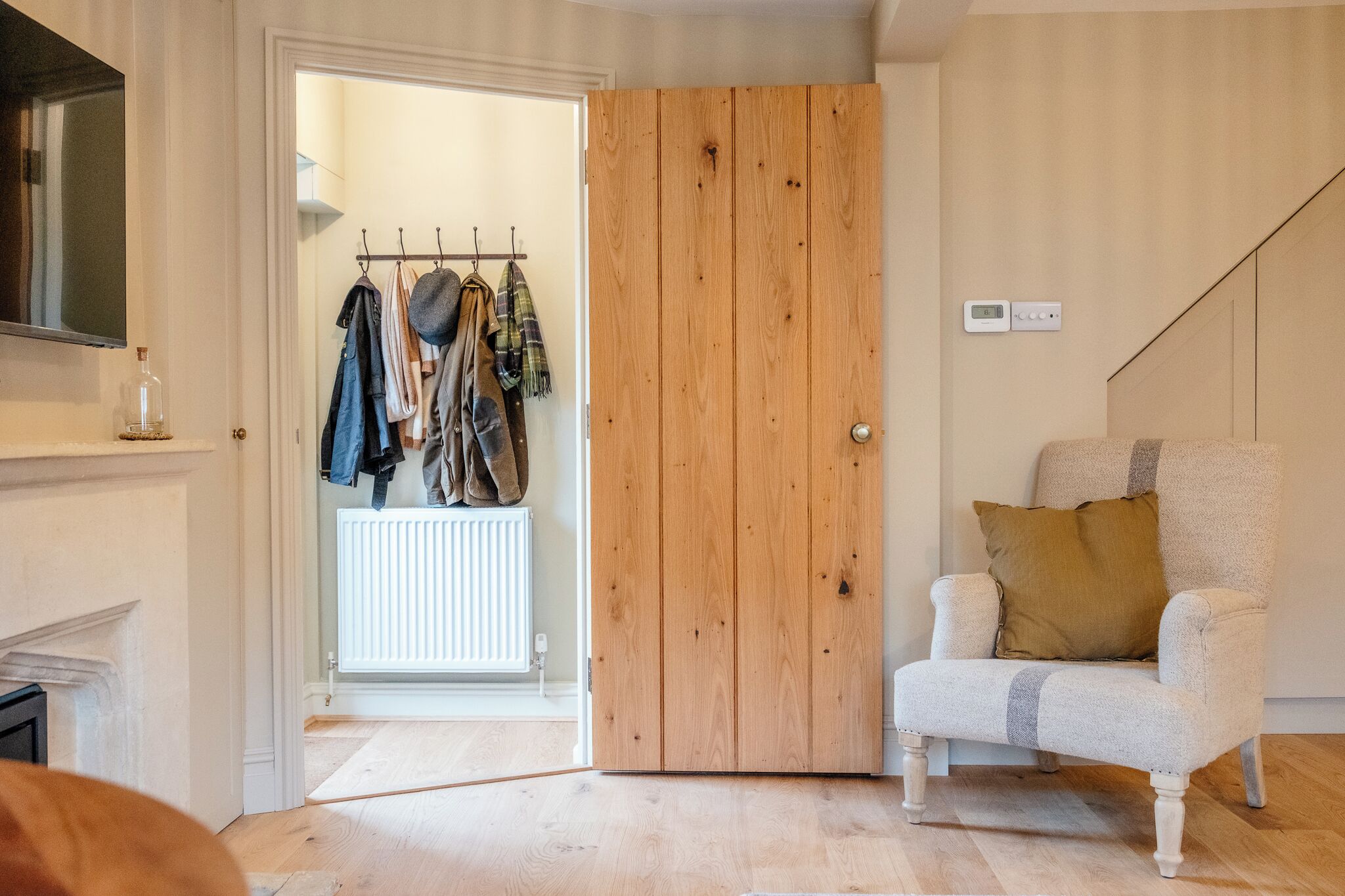 A room with a wooden door, a coat rack with hanging clothes, a white armchair with a cushion, and a thermostat on the wall.