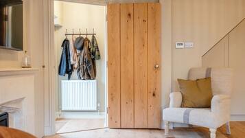 A room with a wooden door, a coat rack with hanging clothes, a white armchair with a cushion, and a thermostat on the wall.