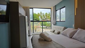 A hotel room with a bed, a TV, and a large window with a view of palm trees. - Nawa Beach Residence 101 by La Playa  (Tamandaré)