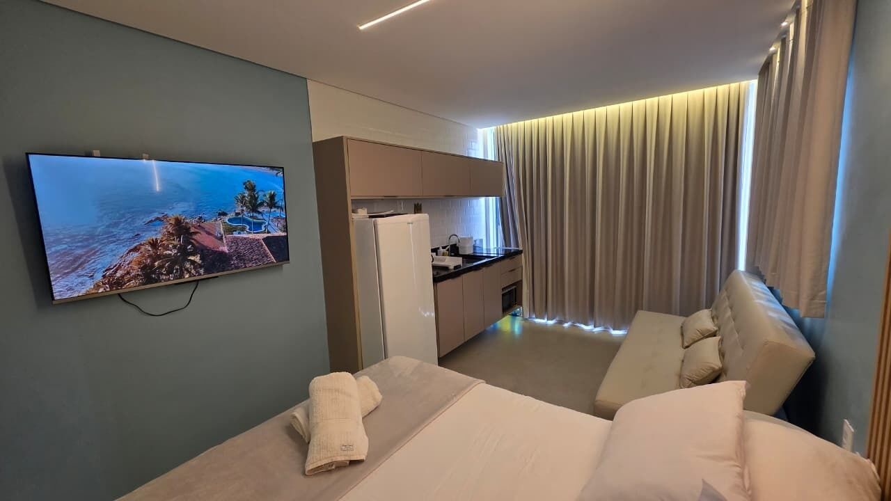 Habitación de hotel con cama, televisor que muestra una vista panorámica, kitchenette y zona de estar con sofá.