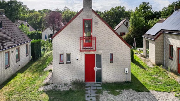 A small, single-story building with a red door and windows, surrounded by a grassy area and other buildings. - 6-person Lake Bungalow in Ewijk (Ewijk)