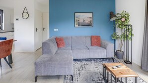 A living room with a grey sectional sofa, a coffee table, a blue wall, a framed picture, a floor lamp, and a potted plant. - 6-person Lake Bungalow in Ewijk (Ewijk)