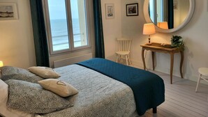 Een slaapkamer met een bed, een stoel, een bureau, een spiegel en uitzicht op de oceaan.