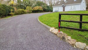 A gravel driveway leading to a white house with a sloped roof, surrounded by greenery and a wooden fence. - Lugdine Lodge (Glengarriff)