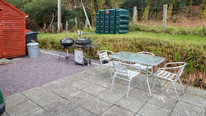 A patio with a table and chairs, a barbecue grill, a trash bin, and a shed. - Lugdine Lodge (Glengarriff)