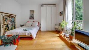 Une chambre à coucher avec un lit, une chaise, un bureau, une télévision et une fenêtre ornée de plantes.