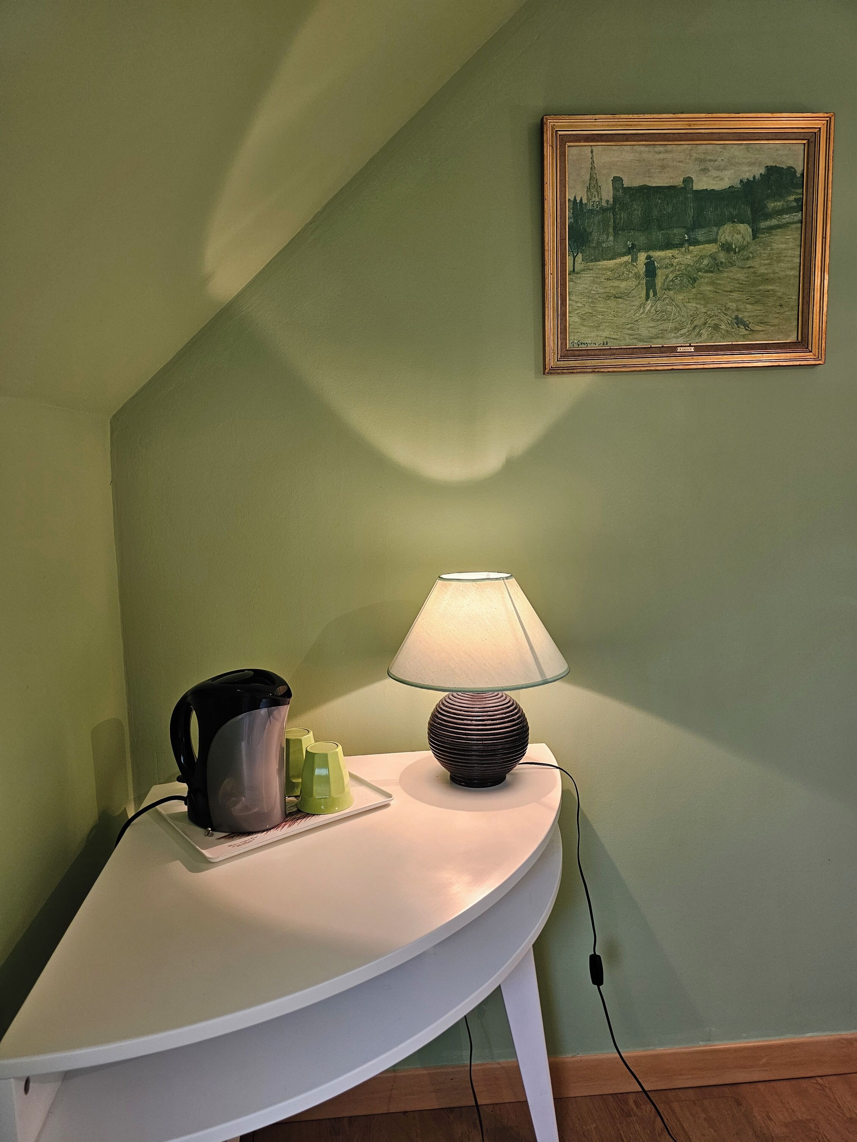 A white bedside table with a lamp, a kettle, and a plate with cups. A framed painting hangs on the wall.