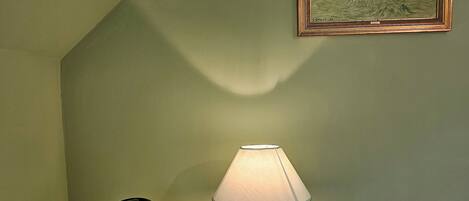 A white bedside table with a lamp, a kettle, and a plate with cups. A framed painting hangs on the wall.