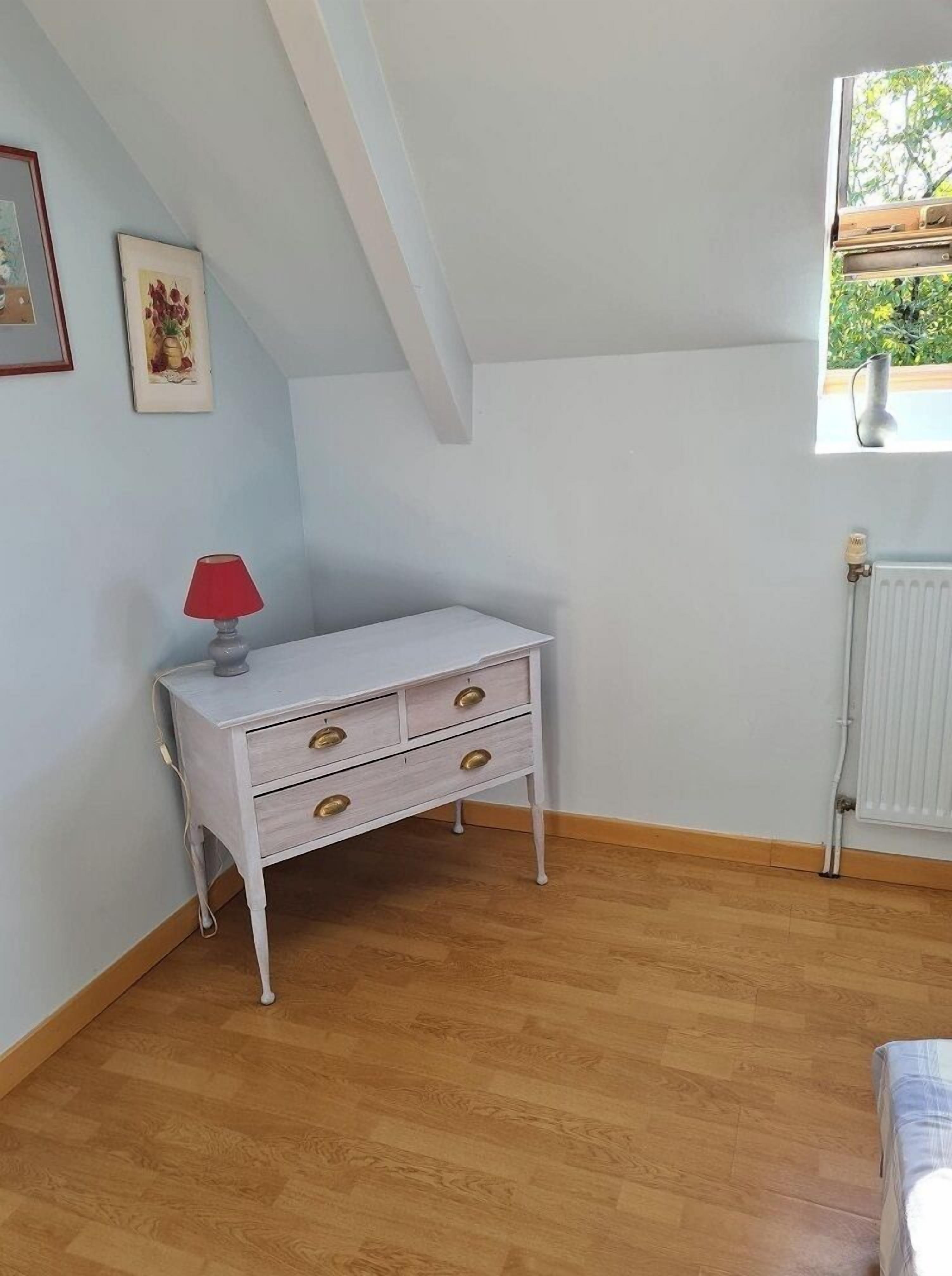 Quarto com cômoda branca, luminária vermelha e janela com vista para o verde.