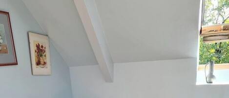 Quarto com cômoda branca, luminária vermelha e janela com vista para o verde.