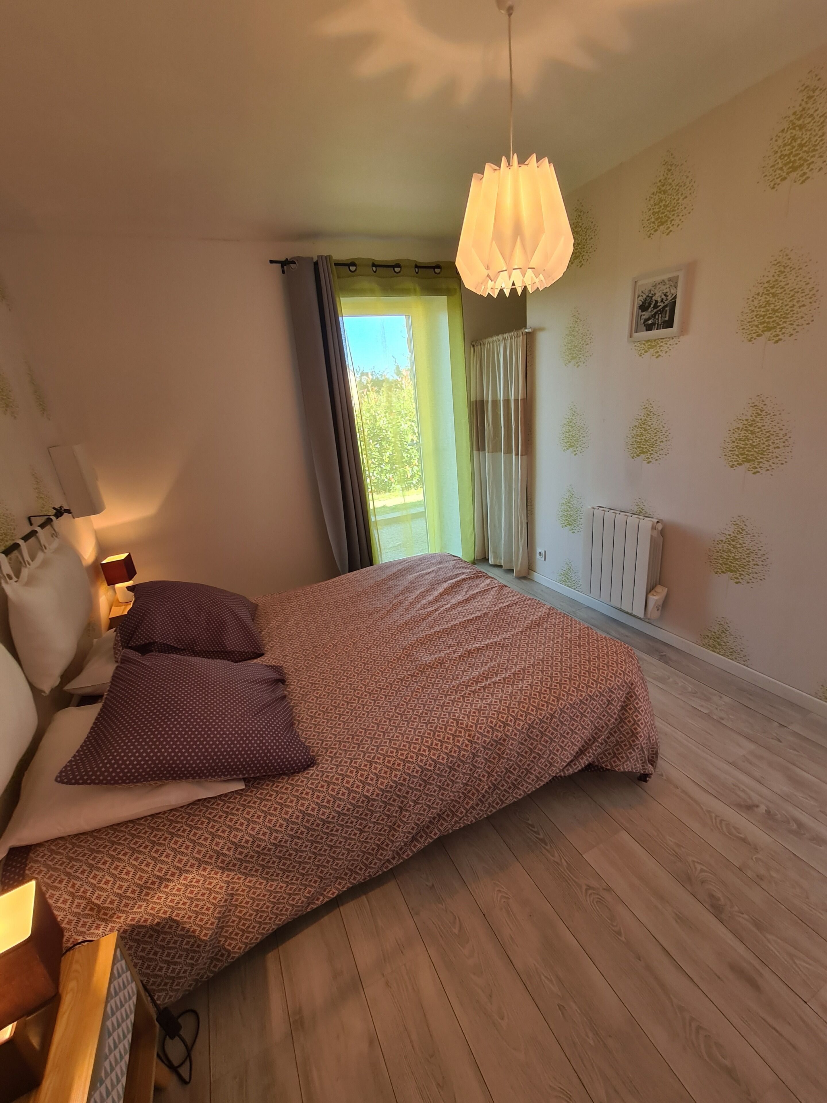 Een slaapkamer met een bed, kussens, een nachtkastje met een lamp, een radiator en een raam met gordijnen.