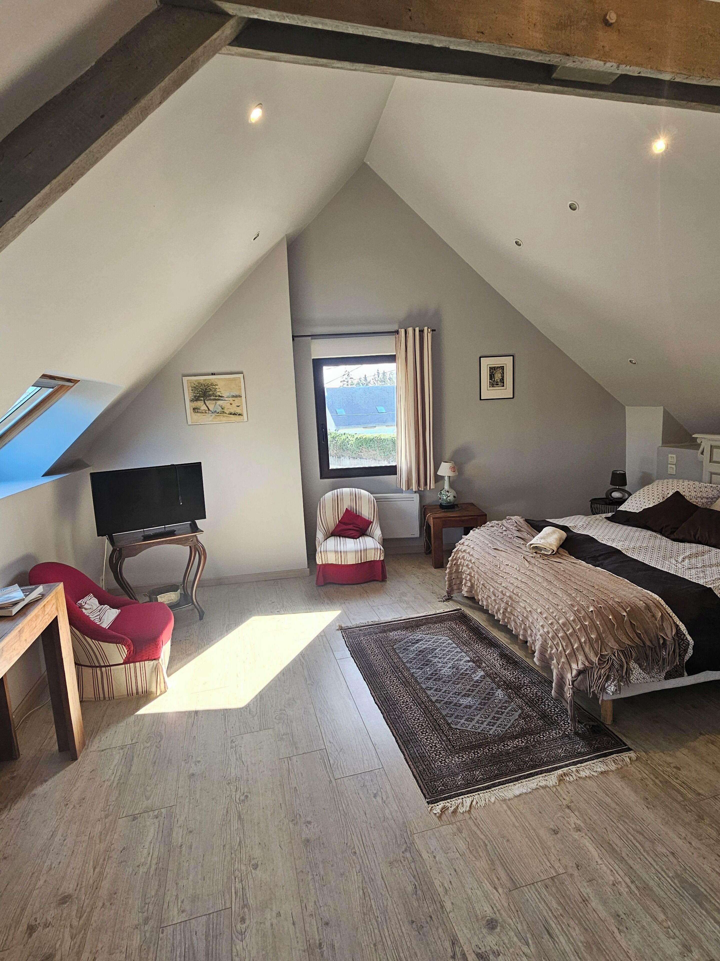 Ein Schlafzimmer mit schrägem Dach, einem Bett, einem Fernseher, einem Sessel, einem Fenster und einem Teppich.