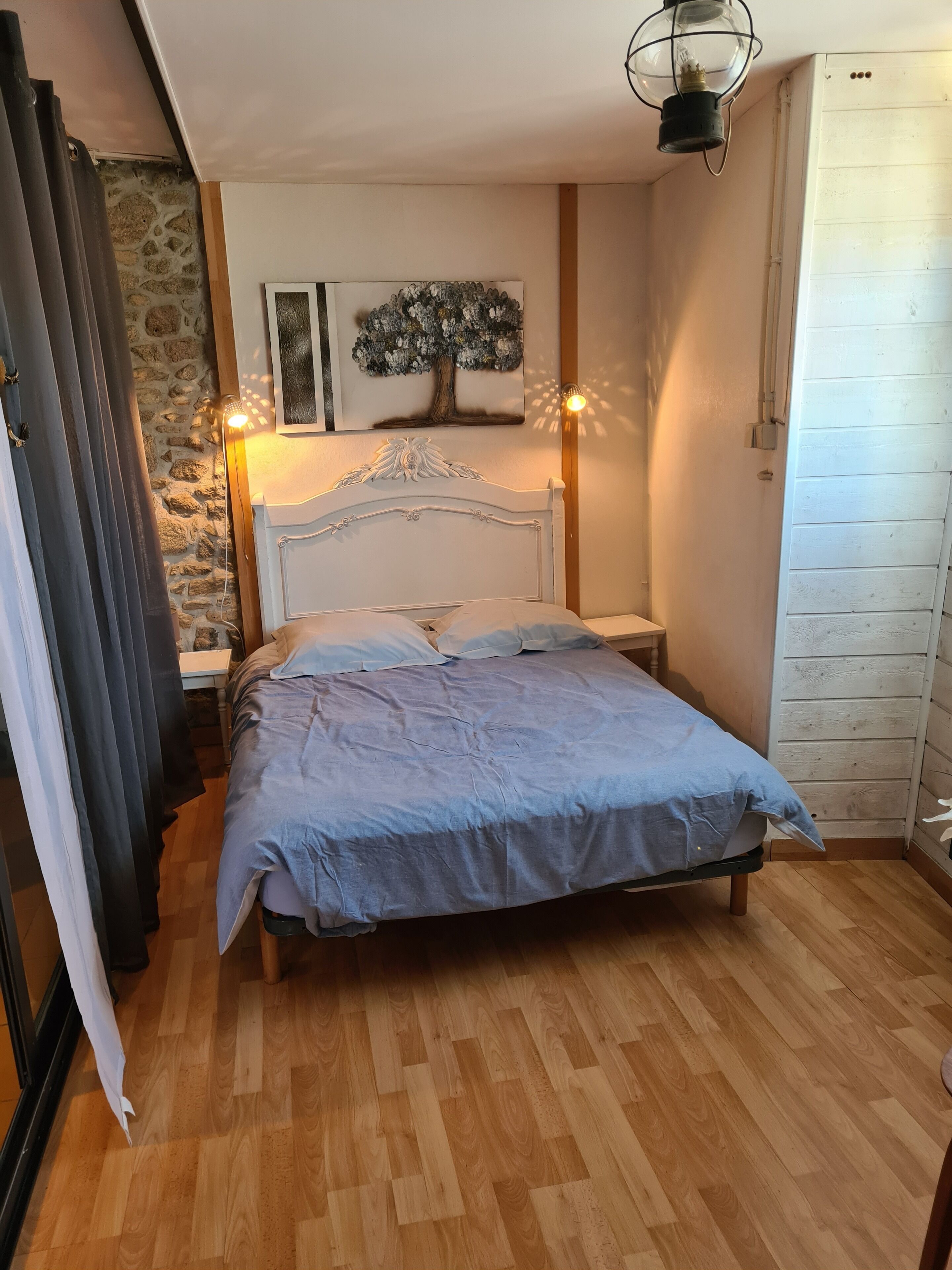 A bedroom with a bed, a nightstand, a lamp, a wall-mounted painting, and a wooden floor.