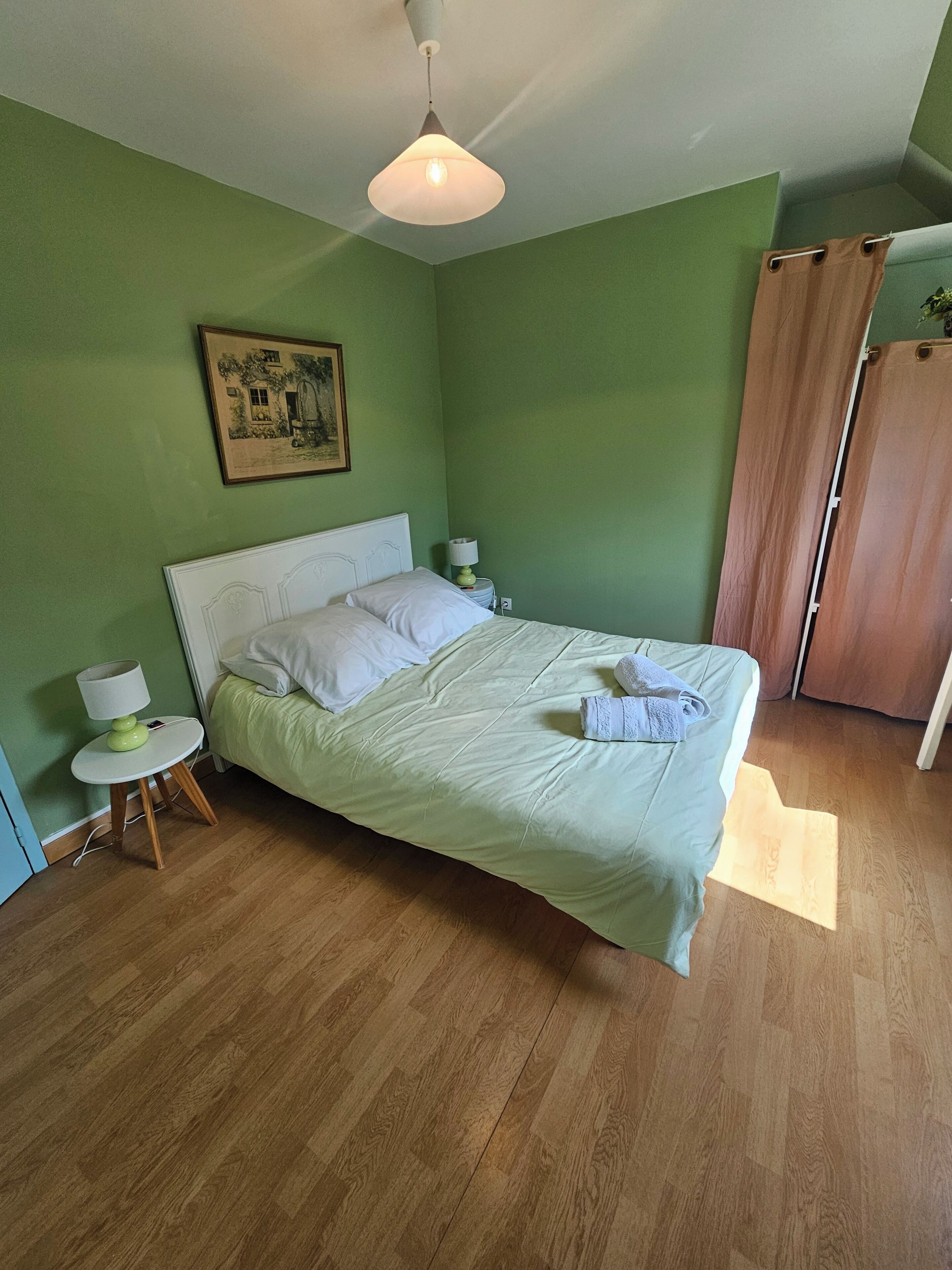 Ein Zimmer mit einem Bett, einem Nachttisch mit Lampe, einem Bild an der Wand und einer Holztür.
