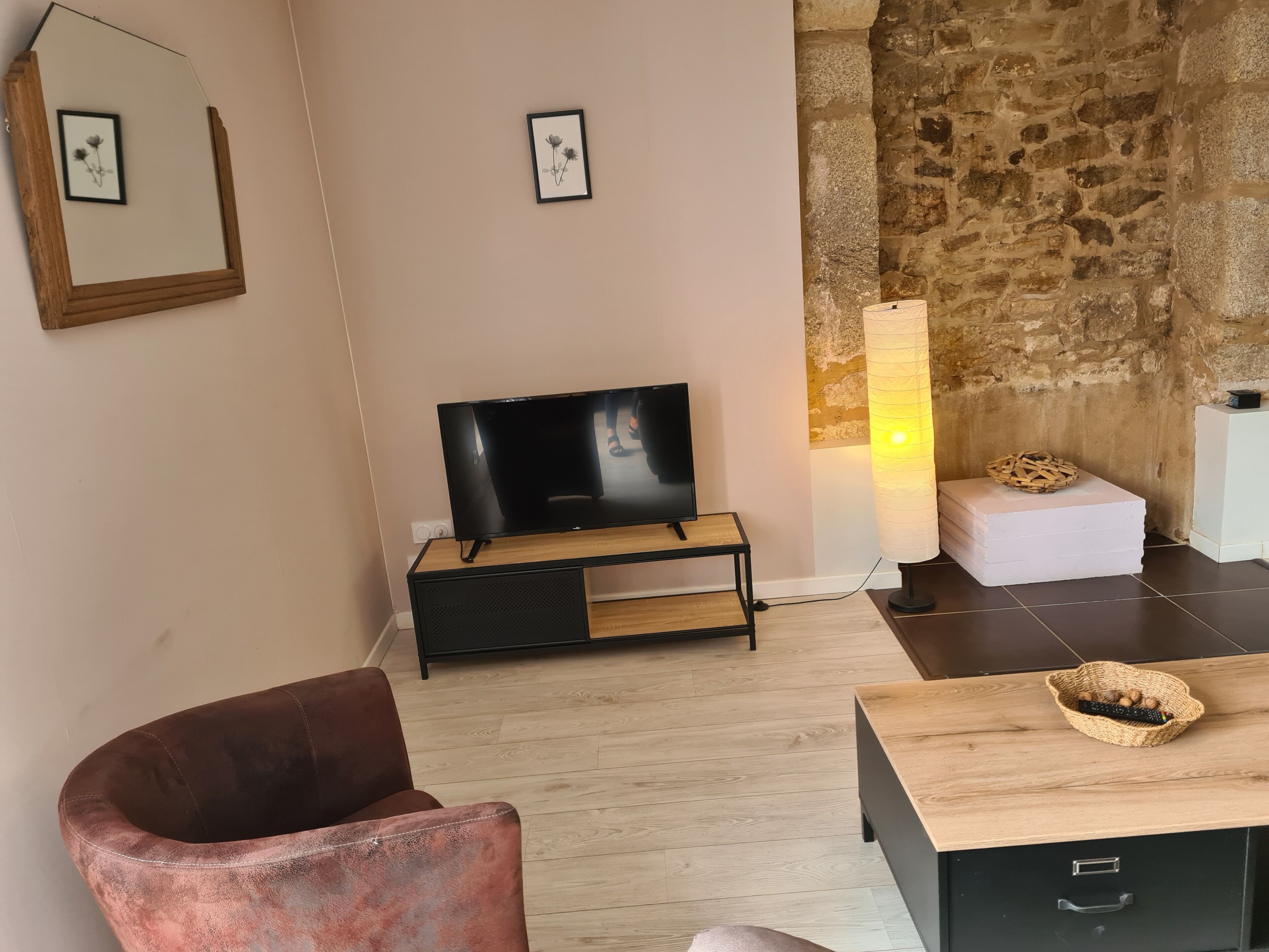 A living room with a flat-screen TV on a wooden stand, a stone wall, a woven basket on a table, and a cushioned chair.