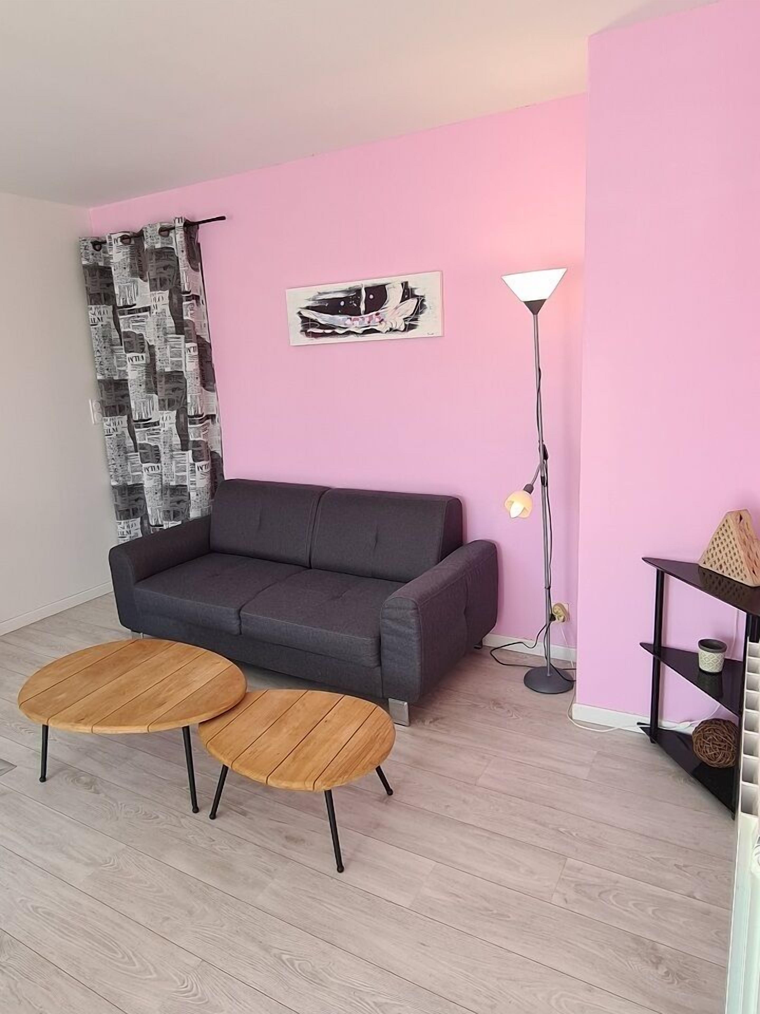 Una sala de estar con un sofá gris, dos mesitas de madera, una lámpara de pie y una pared rosa.