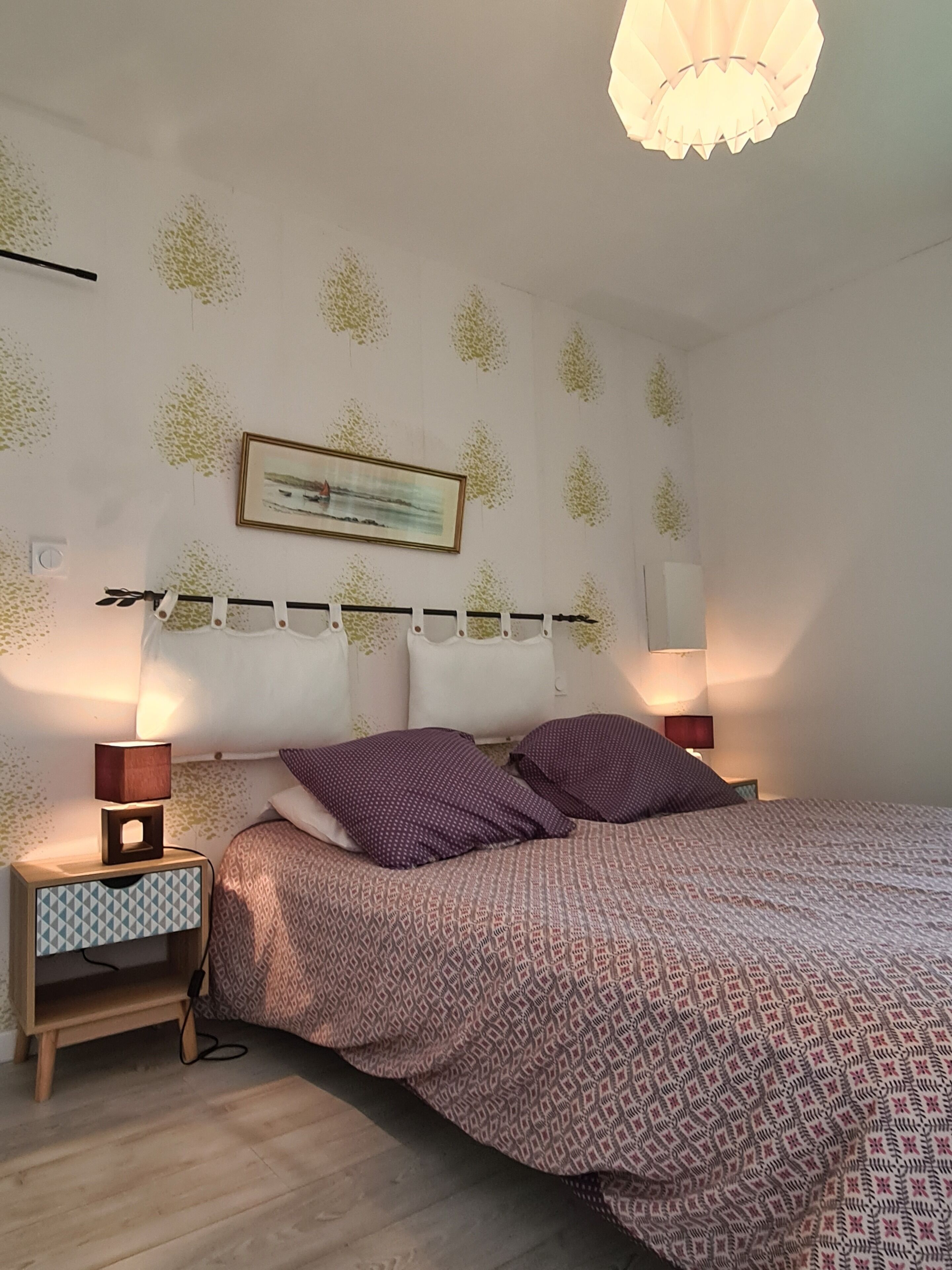 Un dormitorio con una cama, dos mesitas de noche, un velador, un cuadro en la pared y una lámpara del techo.