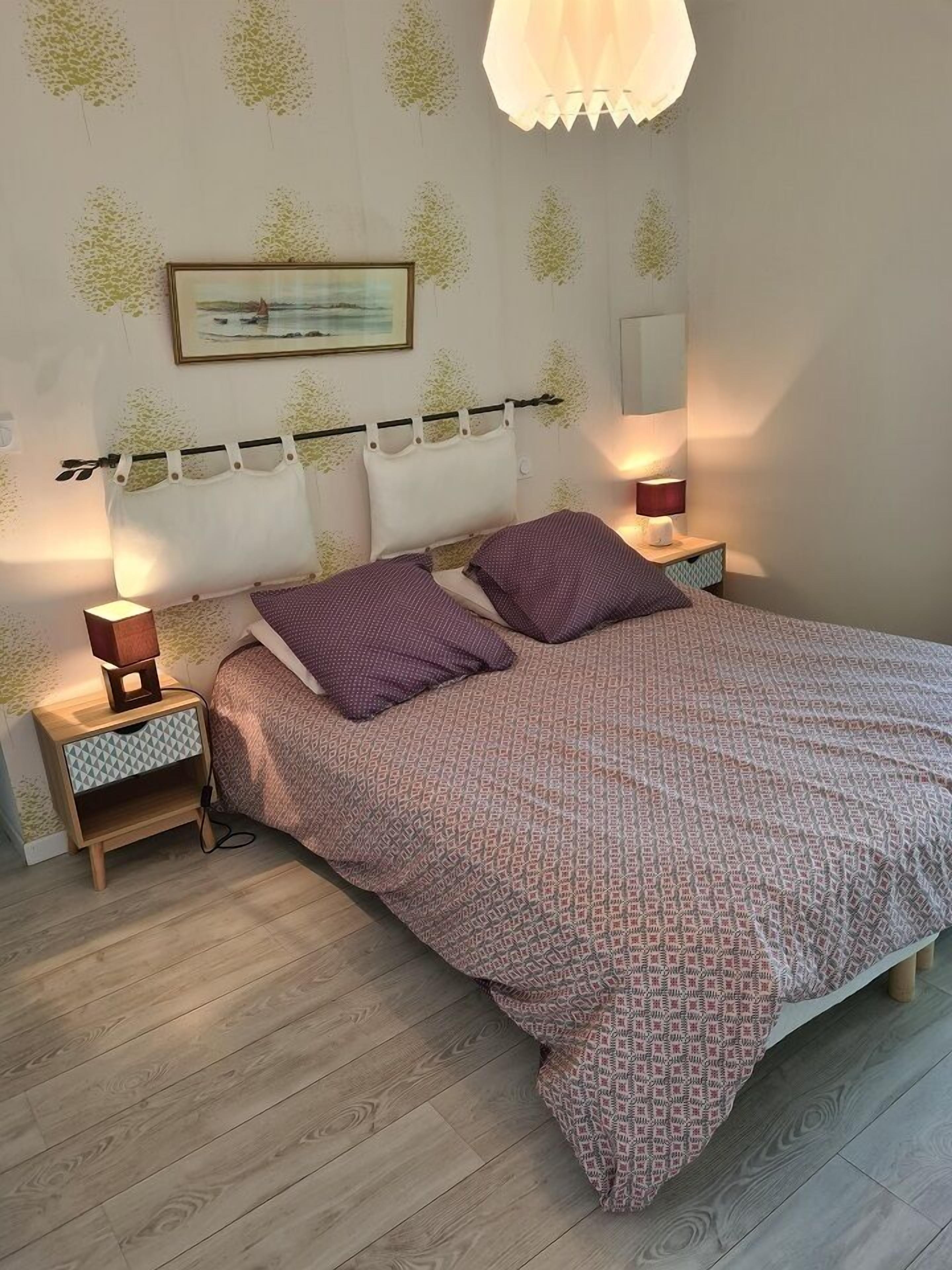 A bedroom with a bed, two bedside tables, a nightstand, a wall-mounted lamp, and a painting.