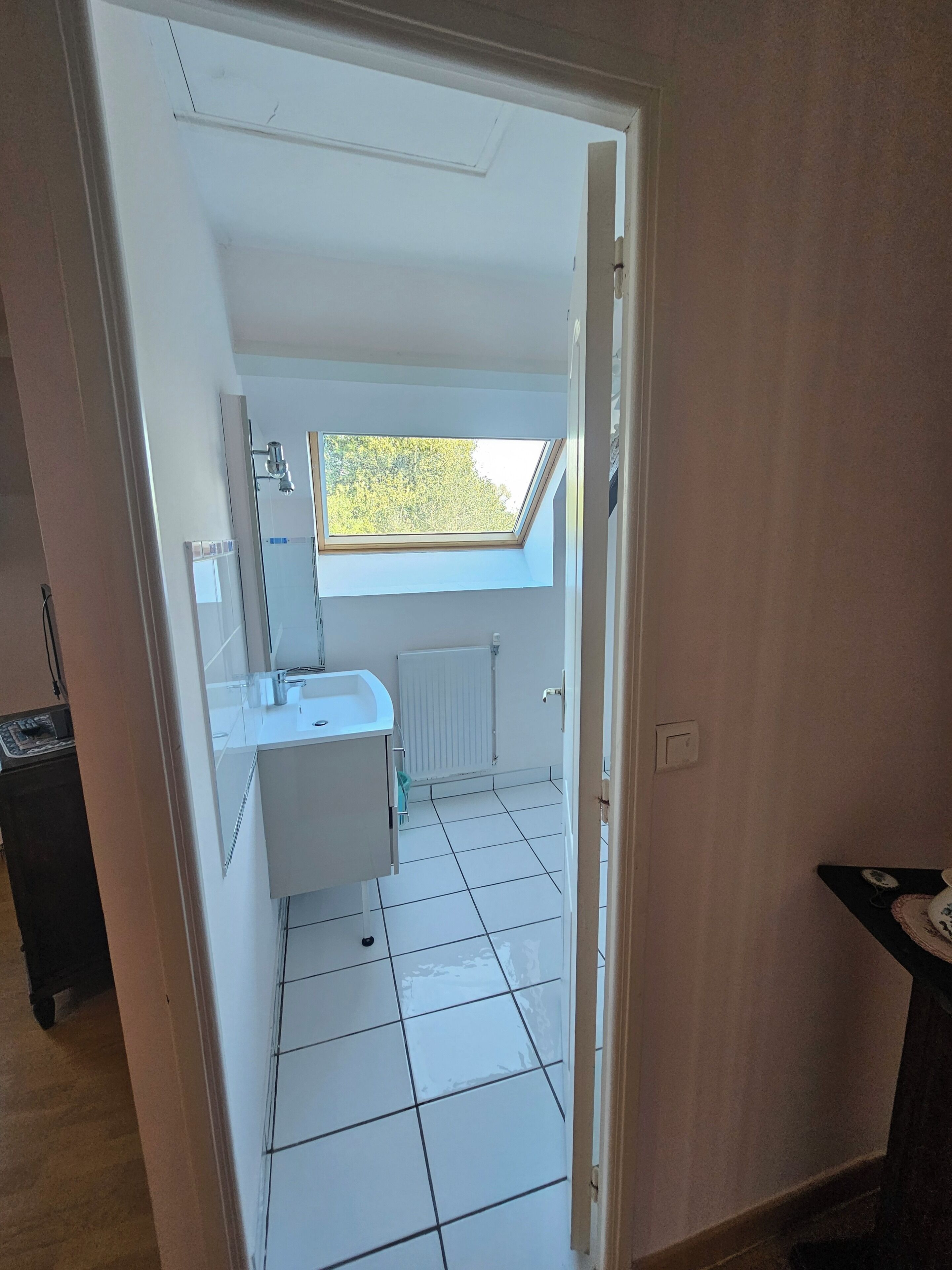 Un baño con tragaluz, azulejos blancos, un lavamanos blanco y un espejo.