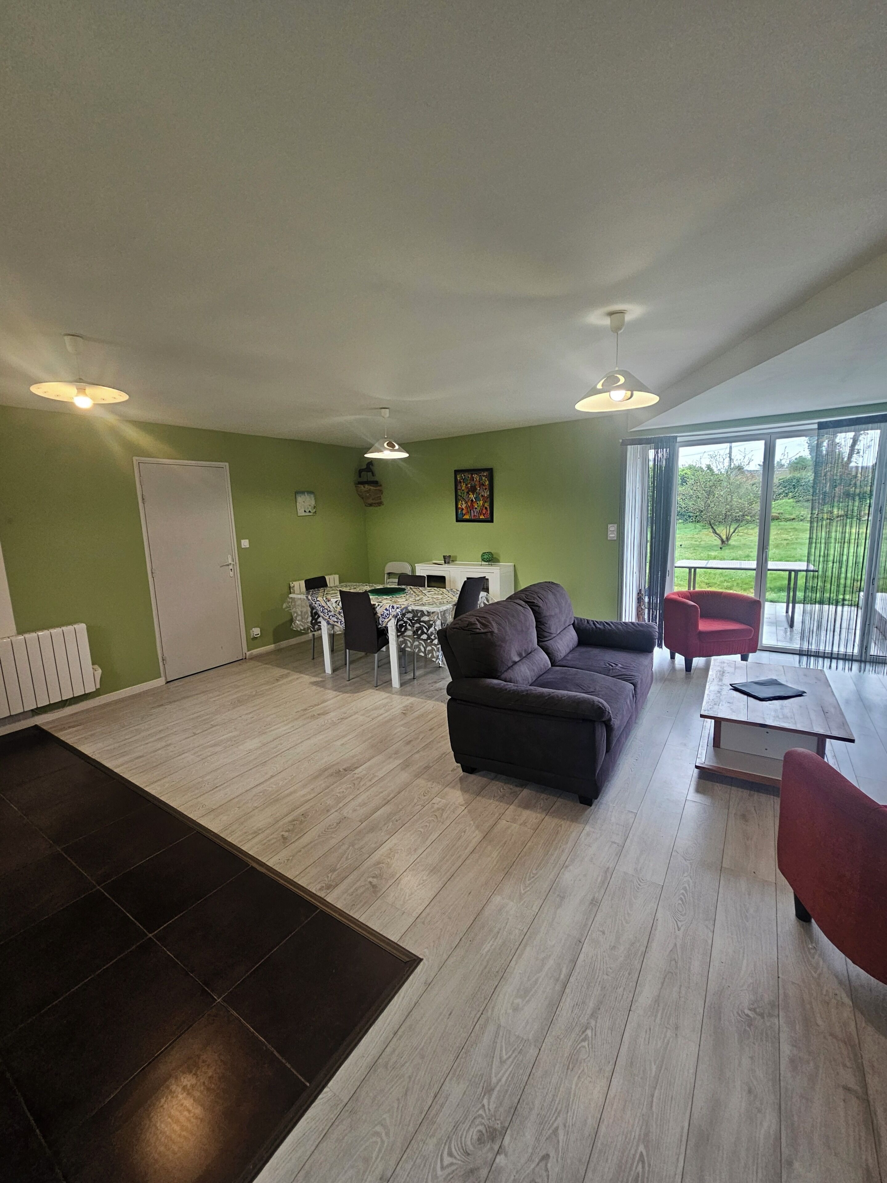 Amplia sala de estar con sofá, una mesa de comedor y sillas, una zona de cocina y un amplio ventanal corredero que da a un jardín.