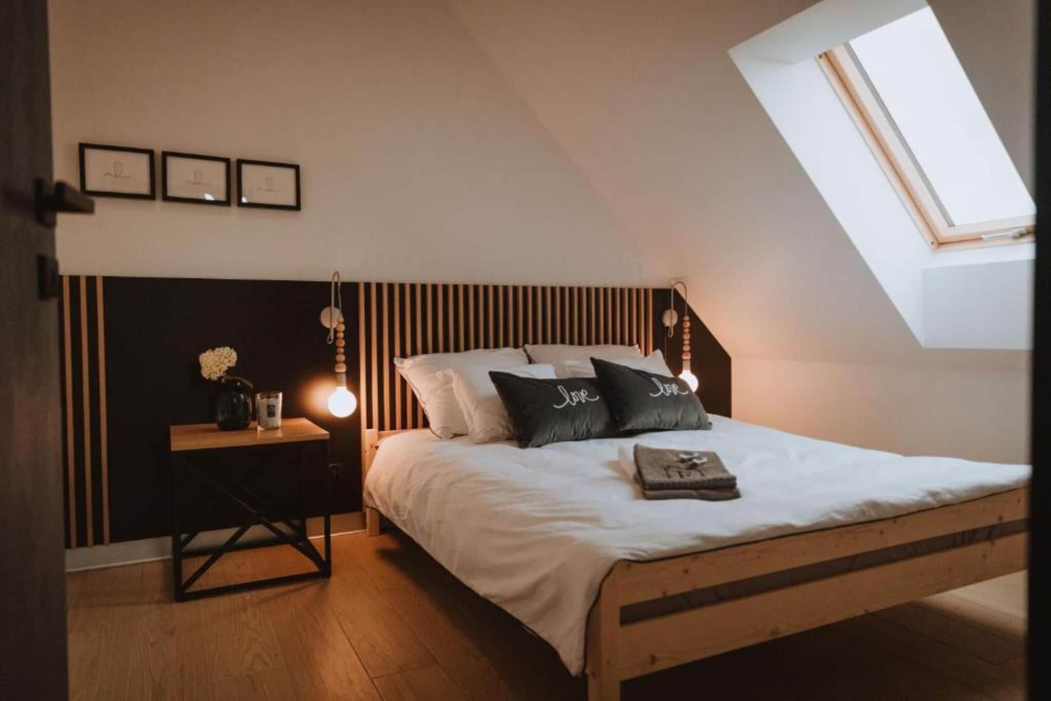 Une chambre avec un plafond en pente, un lit avec une literie blanche, deux oreillers ornés du mot « Amour » brodé, une table de chevet avec une lampe et un petit vase avec des fleurs.
