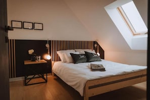 A bedroom with a sloped ceiling, a bed with white bedding, two pillows with 'Love' embroidered, a bedside table with a lamp, and a small vase with flowers.