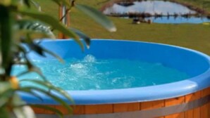 A wooden hot tub with water, surrounded by a wooden deck and a grassy area with a pond and a small bridge. - Large holiday home with a jacuzzi on the terrace (Kościerzyna)